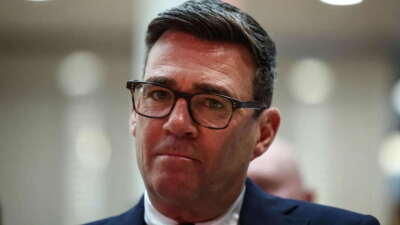 Retrato en plano medio del político británico Andy Burnham usando gafas con montura oscura y traje azul marino.